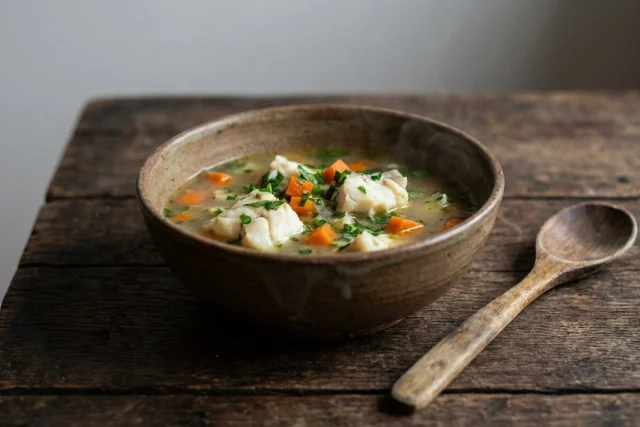 A rustic wooden table setting featuring a ceramic bowl filled with hearty fish broth, chunks of white fish, diced